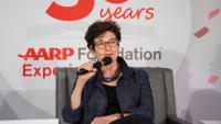 A seated woman speaks into a microphone with the logo AARP Foundation Experience Corps - 30 Years behind her